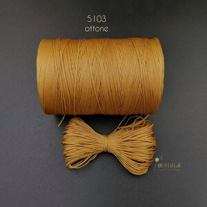 Könnte beinhalten: Eine Spule goldfarbener gewachster Kordel, 5103 ottone, mit einer Schleife aus der Kordel davor.