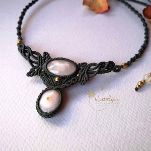Könnte beinhalten: Eine schwarze Makramee-Halskette mit zwei weißen ovalen Steinen und goldenen Akzenten. Die Halskette heißt "Nostalgie Art Jewelry".