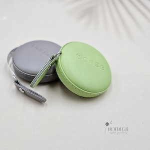 Könnte beinhalten: Zwei runde Ledermaßbänder, eines grau und eines grün. Das grüne Band hat das Wort "tiense" geprägt.