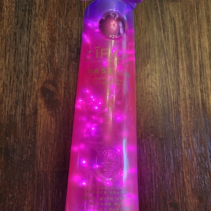 May include: A clear glass bottle of Ciroc Passion Vodka with a purple ribbon around the neck. The bottle is illuminated with pink fairy lights. The label reads "Ciroc Passion, French Grape Vodka, Made with Vodka Infused with Natural Flavors".