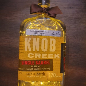 Può includere: Una bottiglia di vetro trasparente con un fiocco marrone e luci a catena all'interno. La bottiglia è etichettata "Knob Creek Single Barrel Reserve Kentucky Straight Bourbon Whiskey" e "Small Batch Aged Nine Years 120 Proof".