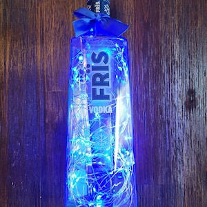 May include: A blue glass bottle of Fris Vodka with a blue ribbon around the neck. The bottle is filled with white fairy lights.