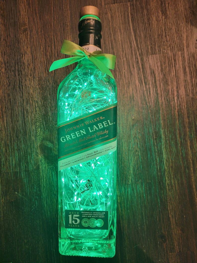 Op de afbeelding: Een doorzichtige glazen fles met groene lampjes erin. De fles is gelabeld met "Johnnie Walker Green Label" en "Blended Scotch Whisky". Het etiket bevat ook de tekst "15 jaar gerijpt".
