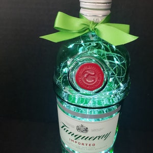 Op de afbeelding: Een groene glazen fles met een zilveren dop en een groene strik. De fles is versierd met witte lampjes en heeft het label "Tanqueray Imported London Dry Gin" erop.