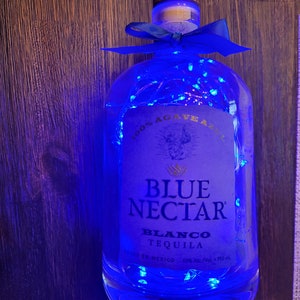 Könnte beinhalten: Eine durchsichtige Glasflasche mit Blue Nectar Blanco Tequila, verziert mit blauen Lichterketten und einem blauen Band. Die Flasche ist mit dem Markennamen und "100% Agave Azul" beschriftet.