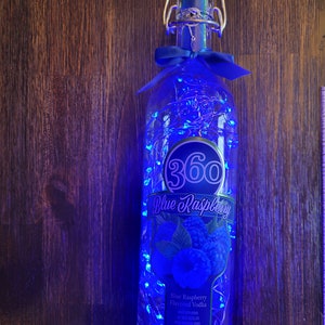 May include: A clear glass bottle containing blue raspberry flavoured vodka. The bottle is decorated with blue LED lights and a blue ribbon. The label reads "360 Blue Raspberry Flavoured Vodka".