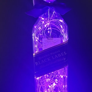 May include: A clear glass bottle with a purple ribbon and string lights inside. The bottle is labeled "Johnnie Walker Black Label Blended Scotch Whisky".
