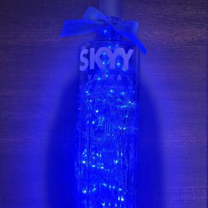 May include: A clear SKYY Vodka bottle filled with blue fairy lights. The bottle has a silver cap and a blue ribbon tied around the neck. The bottle is lit with a blue glow, creating a decorative effect.
