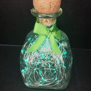 Puede incluir: Una botella de vidrio transparente con un tapón de corcho y una cinta verde. La botella está llena de luces blancas en forma de cuerda que emiten un brillo azul.