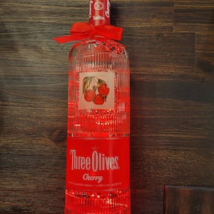 May include: A clear glass bottle of Three Olives Cherry flavoured vodka with a red ribbon tied around the neck. The bottle is illuminated from within, creating a red glow. The label features the brand name "Three Olives" and the flavour "Cherry".
