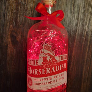 May include: A clear glass bottle with a red ribbon and fairy lights inside. The bottle is labelled "Home Town Horseradish Vodka with Natural Horseradish Flavour 44% Alc/Vol | 88 Proof | 750 ML."