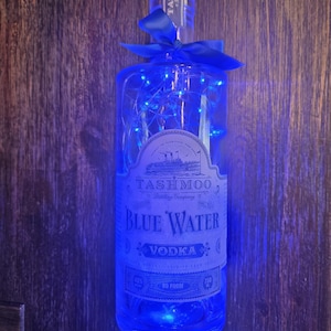 May include: A clear glass bottle of "Blue Water Vodka" with blue string lights and a blue ribbon. The label reads "Tashmoo Distillery Company". The bottle is 750 ml and 80 proof.