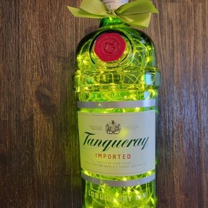 May include: A green glass bottle of Tanqueray London Dry Gin with a silver cap and a green ribbon bow. The bottle is decorated with white string lights. The label reads "Tanqueray Imported, Distilled Four Times, Made from the World's Finest Botanicals, London Dry Gin."