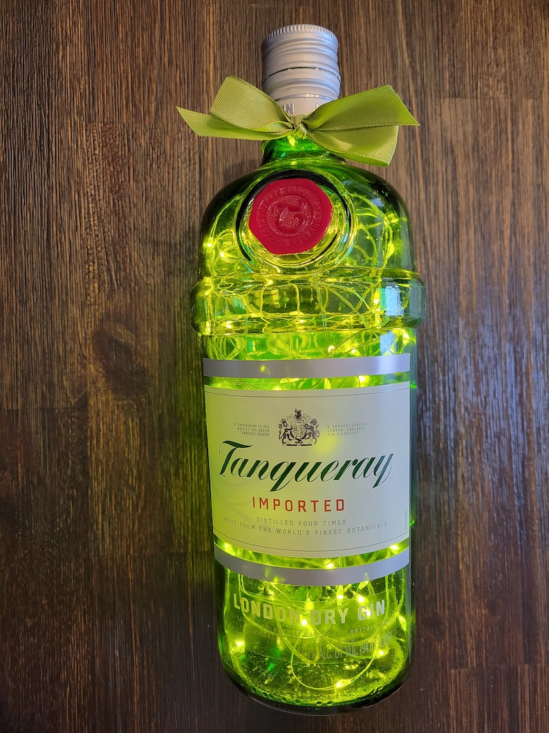 May include: A green glass bottle of Tanqueray London Dry Gin with a silver cap and a green ribbon bow. The bottle is decorated with white string lights. The label reads "Tanqueray Imported, Distilled Four Times, Made from the World's Finest Botanicals, London Dry Gin."