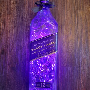 May include: A clear glass bottle with a purple glow from fairy lights inside. The bottle is shaped like a Johnnie Walker Black Label whiskey bottle and has the label text "JOHNNIE WALKER BLACK LABEL BLENDED SCOTCH WHISKY John Walker & Sons AGED 12 YEARS".