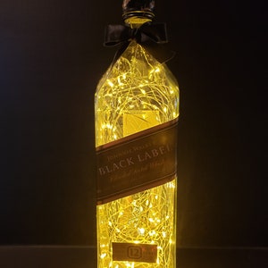 May include: A clear glass bottle with a black bow and string lights inside. The bottle is labeled "JOHNNIE WALKER BLACK LABEL Blended Scotch Whisky 12 YEARS OLD"