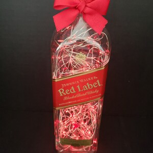May include: A clear glass bottle with a red bow and red string lights inside. The bottle is labeled "Johnnie Walker Red Label Blended Scotch Whisky".