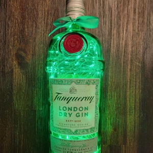 May include: A green glass bottle with a white label and a green ribbon. The bottle is filled with white lights and the label reads "Tanqueray London Dry Gin Est. 1830 Signature Recipe Your Perfectly Balanced Botanicals Produced and Bottled in Great Britain for Charles Tanqueray & Co. London 1L 47.3% alc./vol. England"