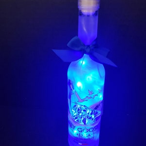 May include: A clear glass bottle with a blue cap and a blue ribbon around the neck. The bottle is filled with white lights and has the words "Grey Goose Vodka" printed on the label.
