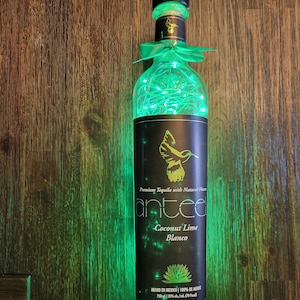 Könnte beinhalten: Eine grüne Glasflasche Anted Coconut Lime Blanco Tequila mit einem grünen Band und Lichterketten darum gewickelt. Die Flasche steht auf einer Holzoberfläche. Das Etikett lautet "Premium Tequila with Natural Flavor" und "Hecho en Mexico | 100% de Agave 750 ml | 35% alc/vol (70 Proof)"