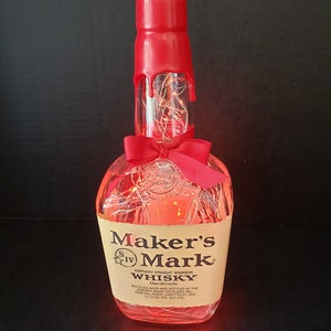 Könnte beinhalten: Eine durchsichtige Glasflasche mit rotem Deckel und einer roten Schleife. Die Flasche ist mit weißen Lichtern gefüllt und trägt ein Etikett mit der Aufschrift "Maker's Mark Kentucky Straight Bourbon Whisky Handmade".