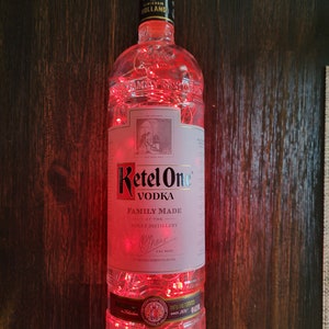 May include: A clear glass bottle of Ketel One Vodka with red fairy lights wrapped around it. The bottle label says "Ketel One Vodka" and "Family Made at the Nolet Distillery".