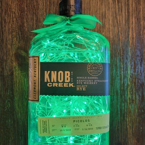 May include: A green glass bottle with a green ribbon and white lights inside. The bottle is labeled "Knob Creek" and "Single Barrel Kentucky Straight Rye Whiskey Select Rye".
