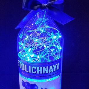 以下が含まれることがあります： 青い妖精ライトで照らされた、Stoli Blueberryウォッカの装飾ボトル。ボトルは金色のキャップ、青いリボン、ブルーベリーのイラストが描かれたラベルが特徴です。ライトは、お祝いの輝く効果を生み出しています。