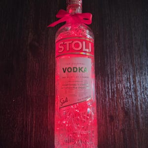 以下が含まれることがあります： 赤いフェアリーライトが入った透明なガラスのストリウォッカボトルと、首に赤いリボンが結ばれています。ボトルにはブランド名と太字の「VODKA」という文字が書かれた白いラベルが付いています。