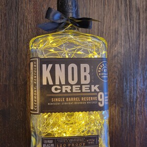 Peut inclure: Une bouteille en verre transparente avec un nœud noir et des lumières de fée à l'intérieur. La bouteille est étiquetée "Knob Creek Single Barrel Reserve 9 Kentucky Straight Bourbon Whiskey 120 Proof".