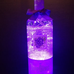 May include: A clear glass bottle with a purple label that says "Three Olives Grape Vodka". The bottle is decorated with a purple ribbon and white fairy lights.