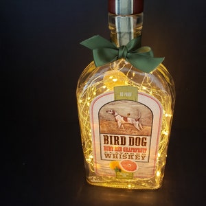 May include: A clear glass bottle with a green bow and string lights inside. The bottle has a label that says "Bird Dog Ruby Red Grapefruit Flavored Whiskey 80 Proof".