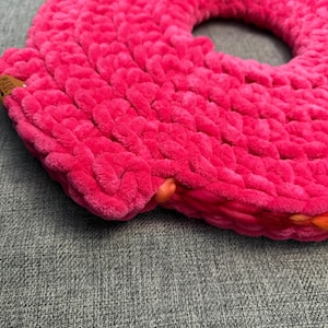 Peut inclure: Un coussin rond rose vif, crocheté, avec un trou au centre. Le coussin est fait de fil épais et moelleux, et les bords sont d'une nuance de rose légèrement plus foncée. Il repose sur une surface grise.