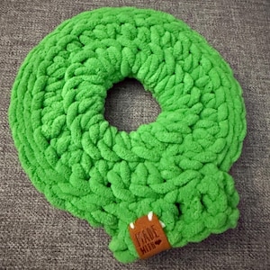 May include: A bright green, handmade, circular crochet item with a central hole. The item is made of thick, textured yarn and features a small brown tag that reads "MADE WITH". The item is likely a decorative pillow or cushion.