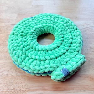 Peut inclure: Un article rond et vert tricoté avec un trou au centre. L'article est fait d'un matériau doux et moelleux et présente un accent bleu sur le bord inférieur. Une petite étiquette indiquant "MADE WITH" est attachée.