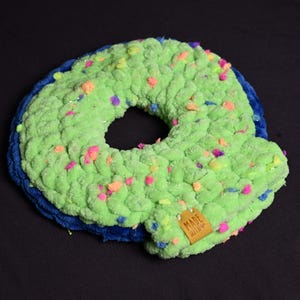 May include: A handmade, plush, green donut-shaped pillow with colorful confetti-like accents. The pillow has a blue base and a small tag that reads "MADE WITH LOVE". The soft, textured design is set against a black background.