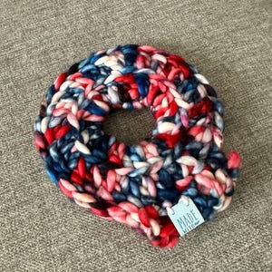 May include: A handmade, chunky knit donut-shaped item in a mix of red, white, and blue yarn. The item has a tag that reads "MADE WITH". The yarn is thick and the stitches are visible, creating a textured appearance.
