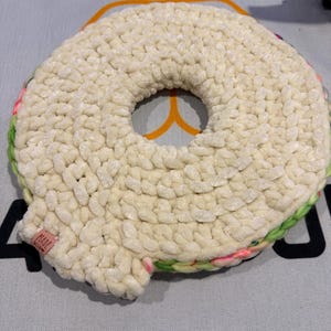 Peut inclure: Un dessous de plat en crochet en forme de donut de couleur crème, avec un trou au centre. Les bords sont ornés de fil vert, rose et blanc. Une petite étiquette indiquant "MADE WITH" est attachée sur le côté.
