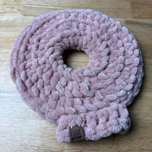 May include: A light pink, donut-shaped hair scrunchie made of a soft, textured material. The scrunchie has a large central hole and a small rectangular tag that reads "MADE WITH". The scrunchie is sitting on a wooden surface.