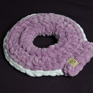 May include: A handmade, donut-shaped pillow in a soft, textured lavender yarn. The pillow has a white border and a small tag that reads "Made with love". The pillow is designed for comfort and support.