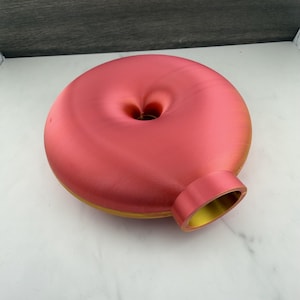Könnte beinhalten: Eine kreisförmige, donut-förmige Vase mit einer glatten, metallischen Oberfläche, die von einem leuchtenden Pink auf der Oberseite zu einem warmen Goldgelb an der Unterseite und im Inneren der Öffnung übergeht, mit zentralem Loch.