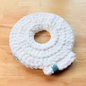 Peut inclure: Un coussin blanc en forme de donut, tricoté en grosse maille, avec un trou central. Le coussin a un aspect doux et texturé et une petite étiquette bleue qui dit "MADE WITH". L'article est placé sur une surface en bois brun clair.