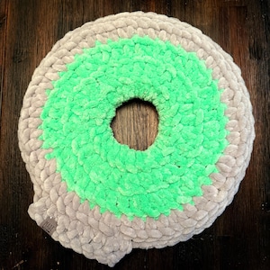 May include: A circular, handmade cushion with a donut shape. The outer ring is a soft grey, and the centre is a vibrant green. The cushion is made of a textured, velvety material. The word "MADE" is visible on a small tag.