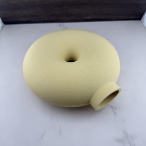 Pode incluir: Um vaso de cerâmica amarelo-pálido em forma de donut com uma textura mate e salpicada. O recipiente apresenta uma abertura circular central e um pequeno bico cilíndrico que se estende a partir do lado, criando um aspeto liso e uniforme.