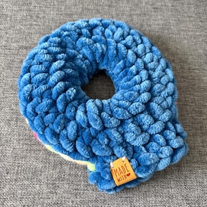 May include: A blue, handmade scrunchie with a plush, textured appearance. The scrunchie has a donut shape with a hole in the center. A small, rectangular tag with the words "MADE WITH" is attached to the scrunchie.