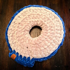 May include: A handmade, circular pet bed in a donut shape. The bed features a soft, plush texture with a light beige center and a vibrant blue border. A small tag with the words "MADE WITH" is attached to the blue border. The bed is designed for comfort.
