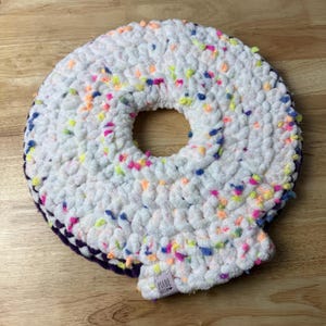 May include: A circular, donut-shaped crochet pillow made of white yarn with multi-colored flecks of pink, blue, yellow, and orange. The underside is a solid dark purple color. A small white fabric tag reads MADE WITH LOVE in black text.