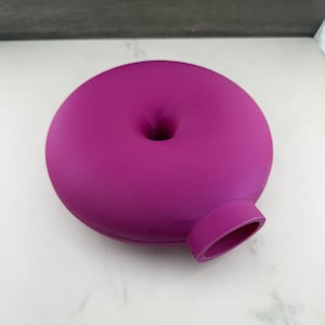 Puede incluir: Un florero circular de color magenta vibrante con forma de rosquilla y acabado mate suave. El objeto presenta una abertura central circular y un pequeño cuello cilíndrico que se extiende desde el lateral, diseñado para sostener un tallo floral.