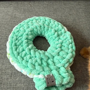 May include: A handmade, mint green crocheted scarf with a circular design. The scarf is made of a soft, chunky yarn and features a small tag that reads "MADE WITH". The scarf is displayed on a gray surface.