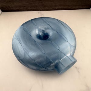 May include: A translucent, metallic blue, 3D-printed vase with a rounded, flattened, donut-like shape. The surface features a repeating pattern of curved, overlapping lines and a central circular opening, with a short, cylindrical neck extending outward.